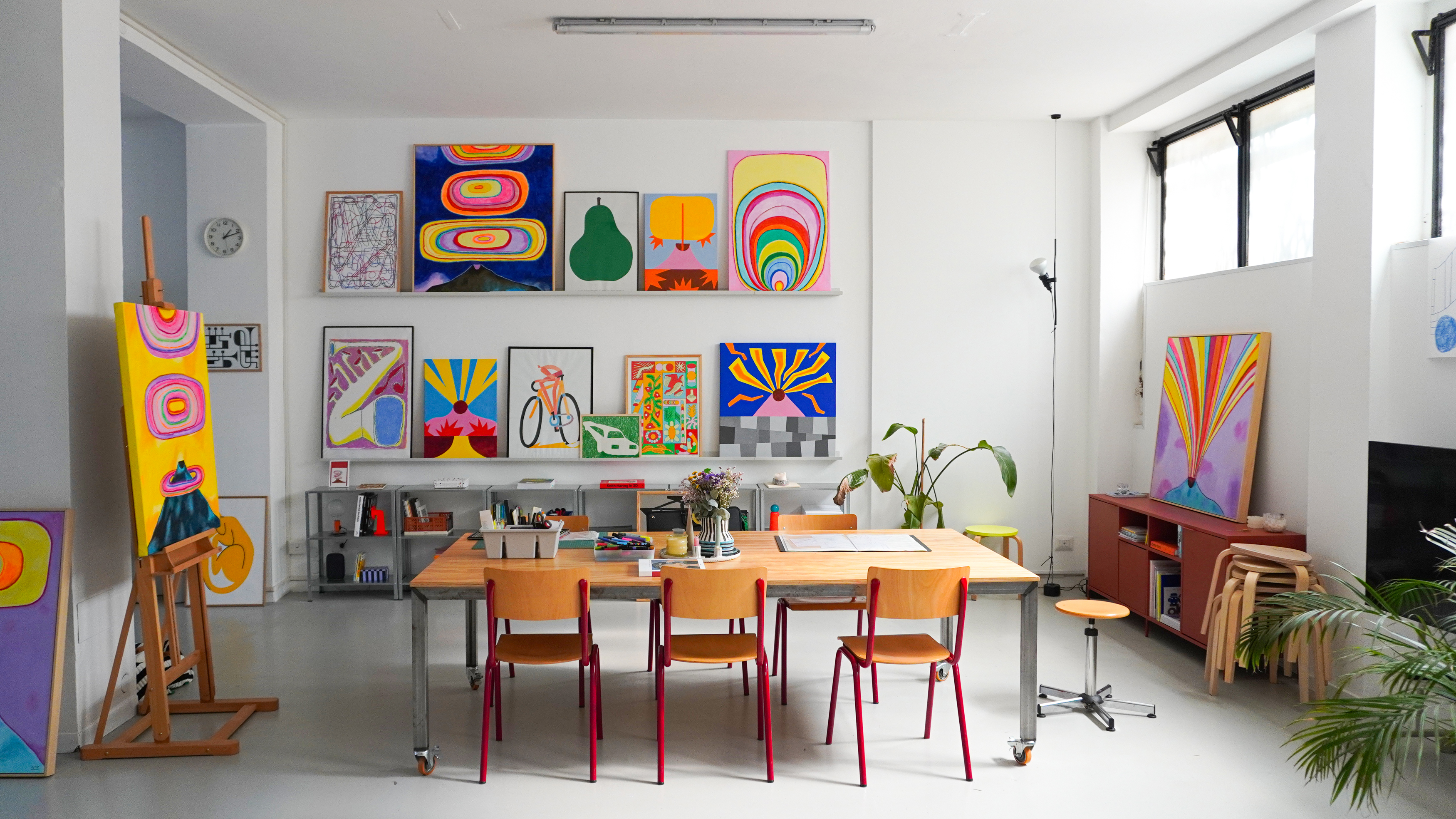 Molto studio interior with colorful paintings, a wooden table and red chairs